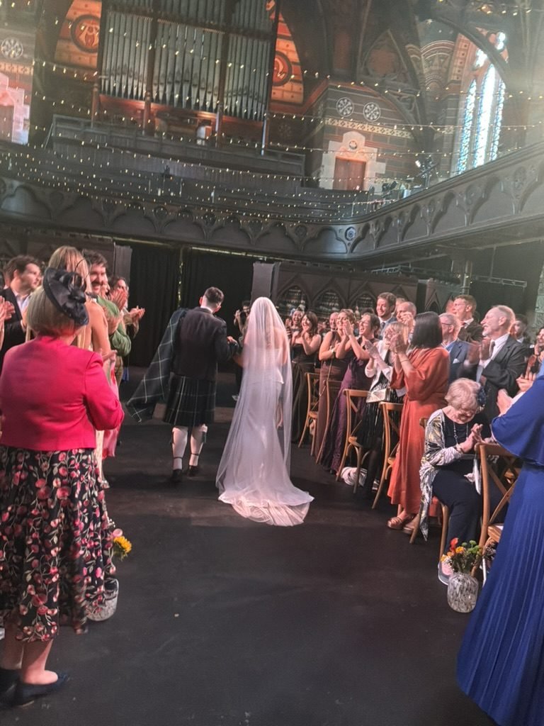 Martha and Gavin walking back down the aisle at Cottiers after their ceremony, guests applauding, the painted organ pipes and fairy-lit gallery behind them