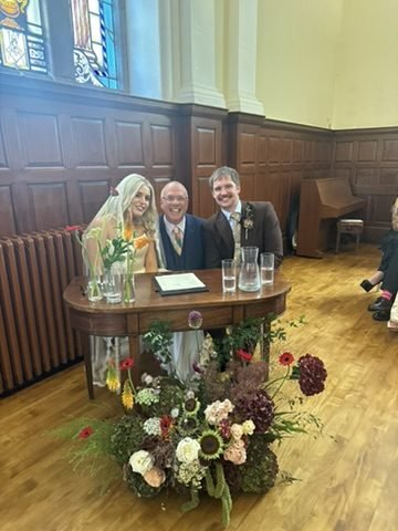 Nadia and Rob seated at the signing table in the Maxwell Hall at Pollokshields Burgh Hall with celebrant John Paul between them, wood panelling and a stained glass window behind and a big autumn flower arrangement in front