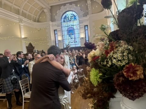 Nadia and Rob kissing in the aisle of the Maxwell Hall at Pollokshields Burgh Hall as guests stand and applaud, fairy lights strung across the hall and a tall stained glass window at the far end