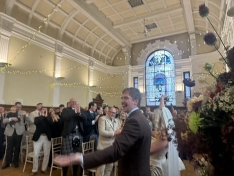 Rob grinning with his arm out as he and Nadia head back up the aisle of the Maxwell Hall as newlyweds, guests on their feet clapping beneath fairy lights and the stained glass window