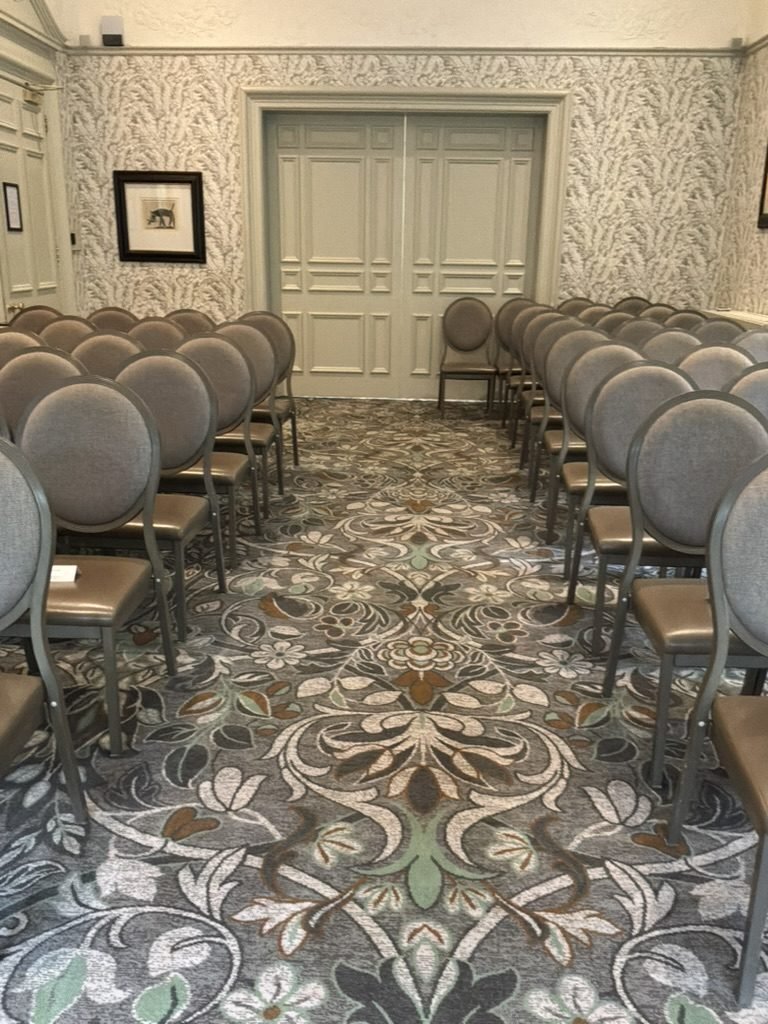 A ceremony room at One Devonshire Gardens set for a wedding, rows of grey upholstered chairs either side of an aisle of patterned carpet, leaf-patterned wallpaper and tall panelled double doors at the far end