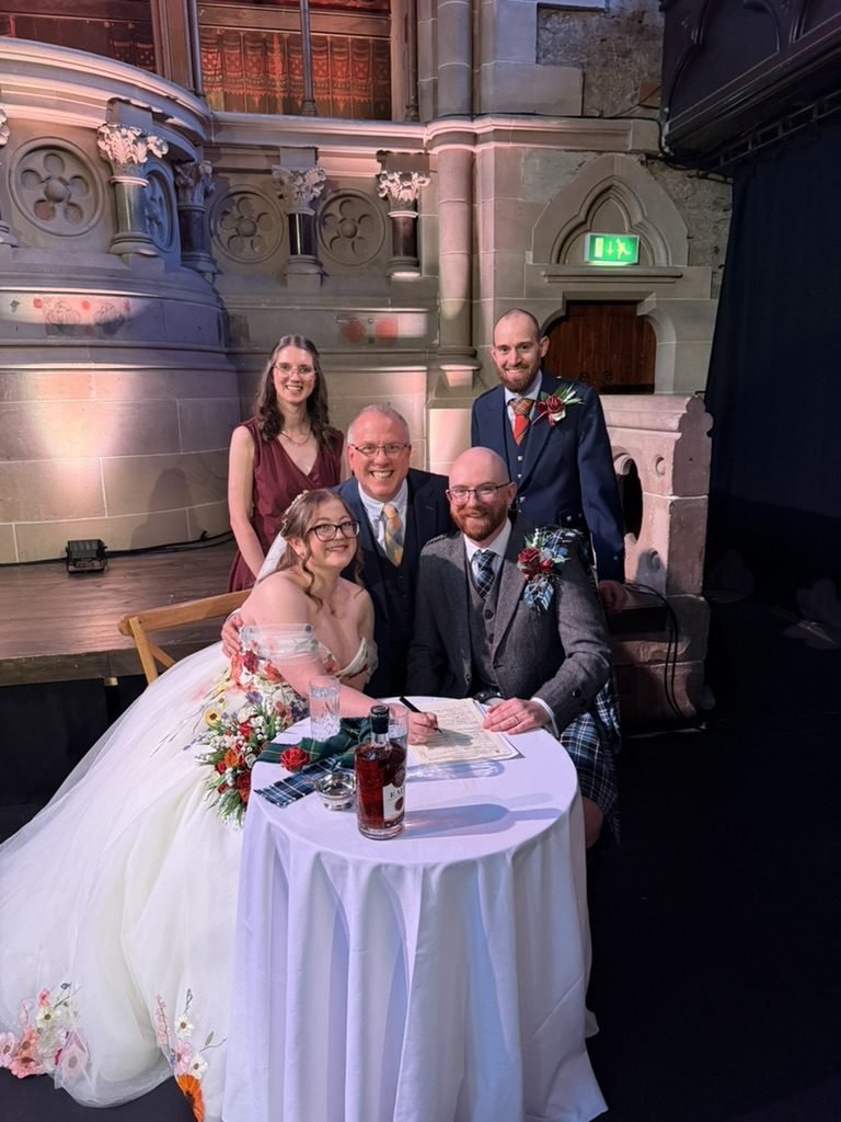 Rachel and Andrew at the signing table at Cottiers with their maid of honour, best man and celebrant John Paul Fitzpatrick, the carved stone of the old church behind them