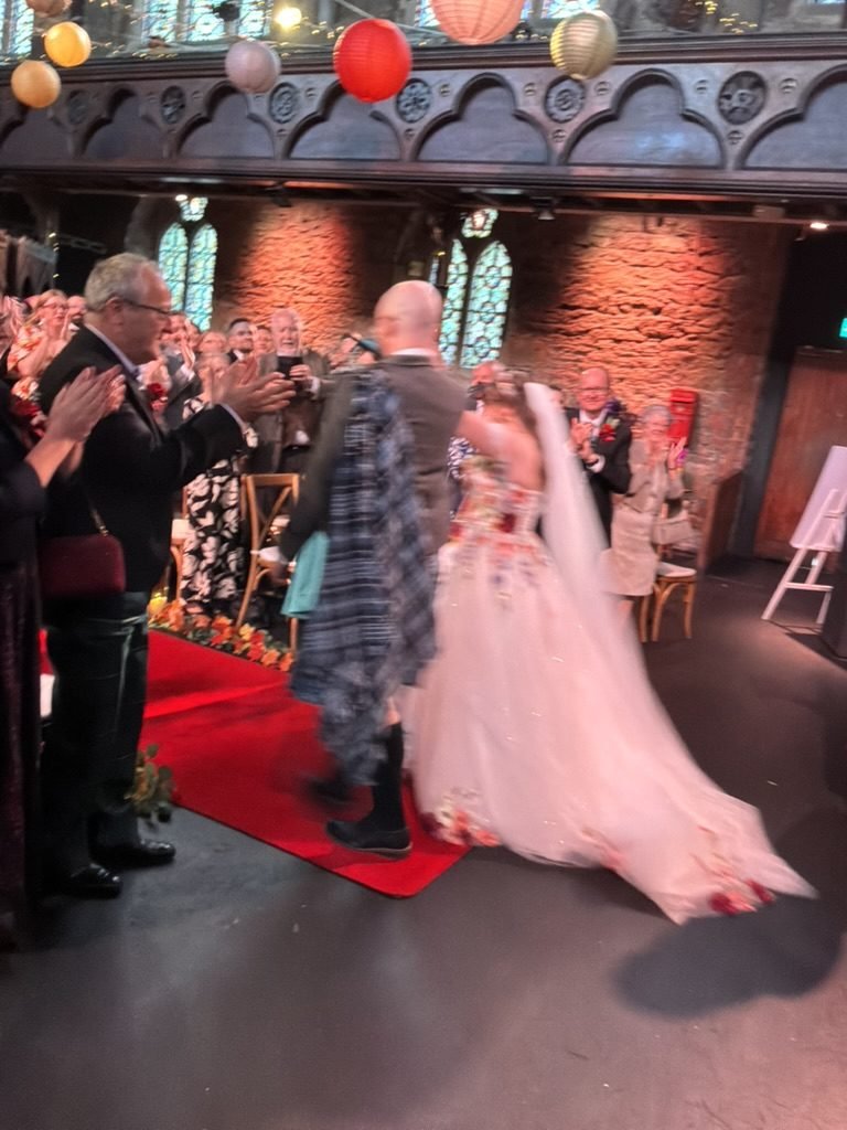 Rachel and Andrew walking back up the red aisle at Cottiers as newlyweds, Andrew in a tartan kilt and Rachel in a gown embroidered with flowers, guests applauding on both sides