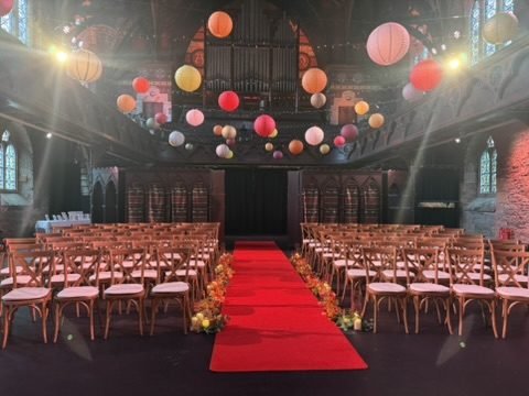The ceremony hall at Cottiers set for the wedding: rows of wooden chairs, a red aisle lined with orange and red flowers, and paper lanterns hanging under the organ pipes