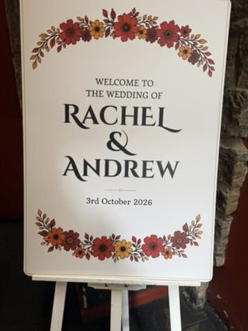 A welcome sign on an easel reading Welcome to the wedding of Rachel and Andrew, 3rd October 2026, framed with red and orange flowers
