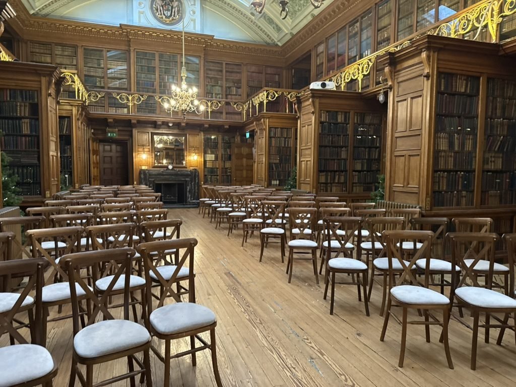 The New Library at the Royal College of Physicians of Edinburgh set for a December wedding, rows of wooden chairs on a timber floor between tall oak bookcases, a gallery with gilded railings above, a chandelier and a dark marble fireplace at the far end and a Christmas tree at the edge