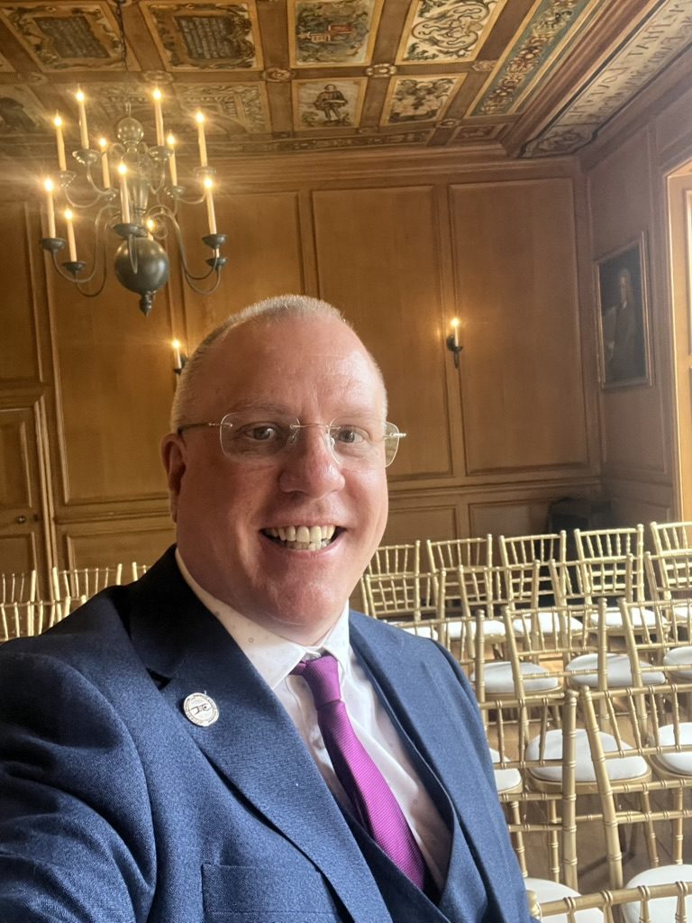 Celebrant John Paul alone in the empty ceremony room at Riddles Court before guests arrive, ornate painted ceiling, chandelier and rows of gold chiavari chairs