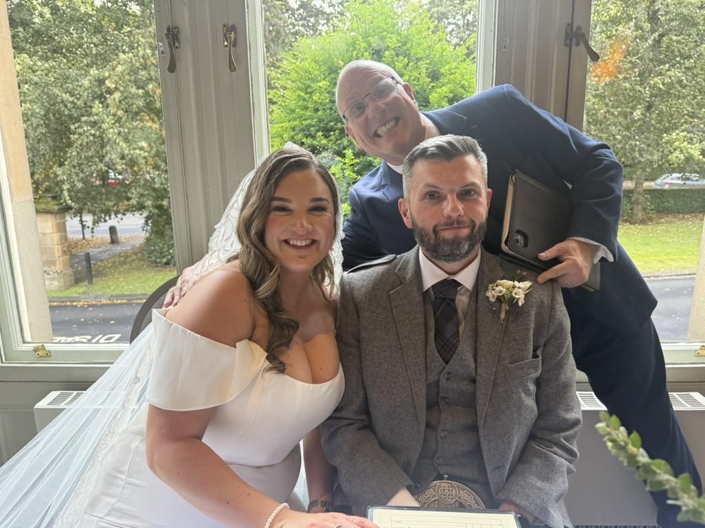 Rhona and Stephen smiling at the signing table in a big bay window at One Devonshire Gardens, with celebrant John Paul leaning in behind them and green trees outside