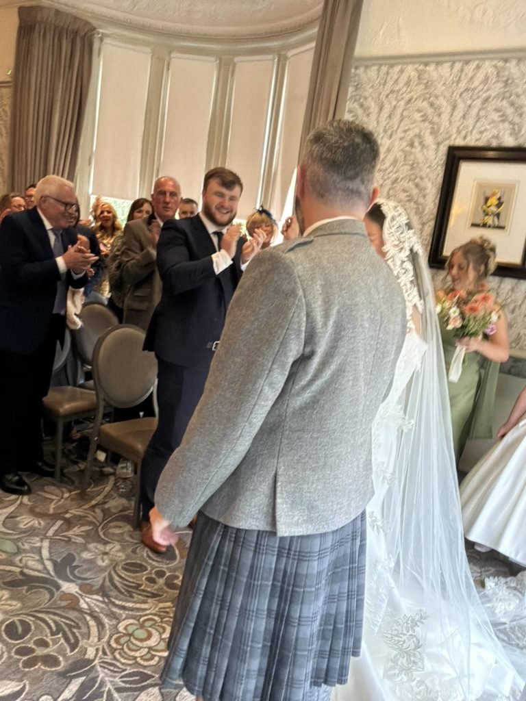 Stephen in a grey tweed jacket and kilt beside Rhona in her long lace-edged veil as guests stand and applaud them in the ceremony room at One Devonshire Gardens
