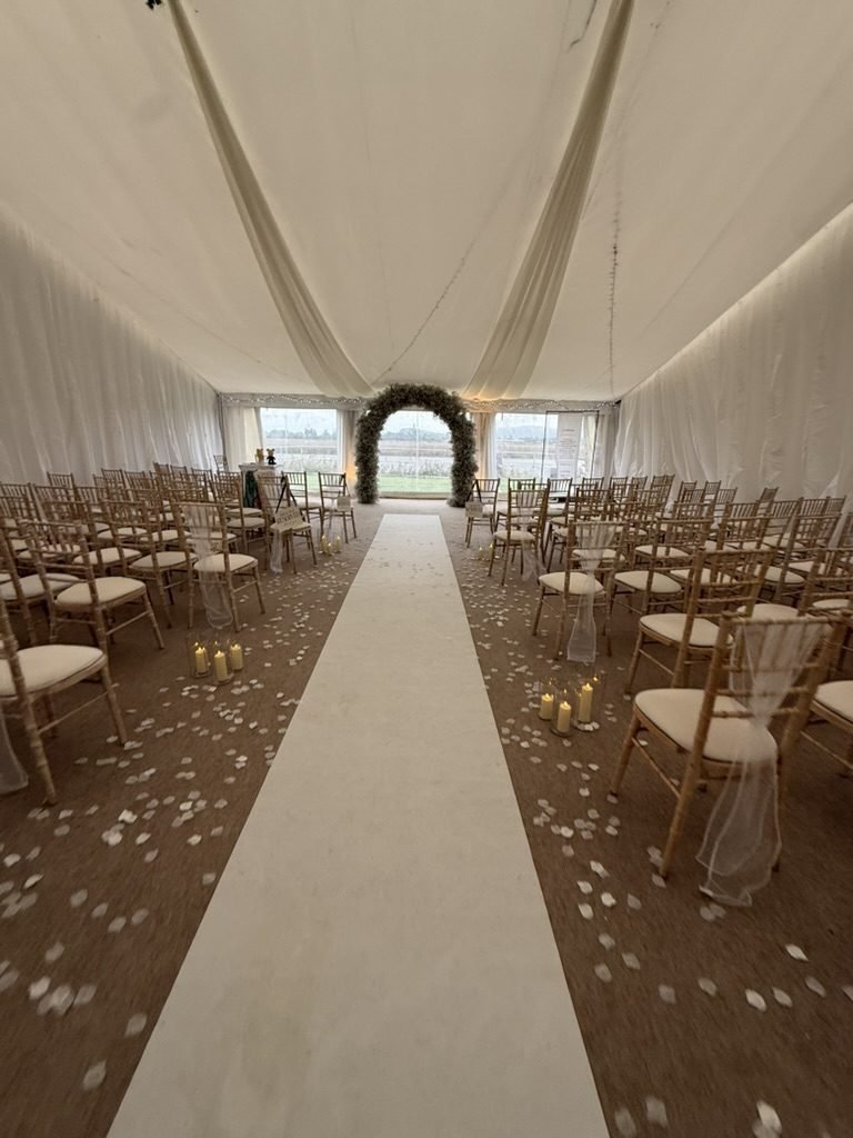 The marquee at The Riviera at Lux set for a humanist wedding: white drapes across the ceiling, gold chairs either side of a long white aisle scattered with petals and candles, and an arch of white flowers in front of the windows at the far end