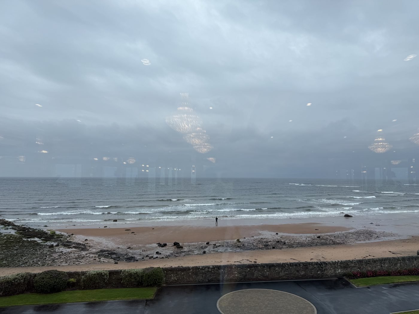 The view from inside the Firth Pavilion at Seamill Hydro down onto the beach: the sea wall and lawn in the foreground, rocks and sand below, and the Firth of Clyde stretching out under a grey sky