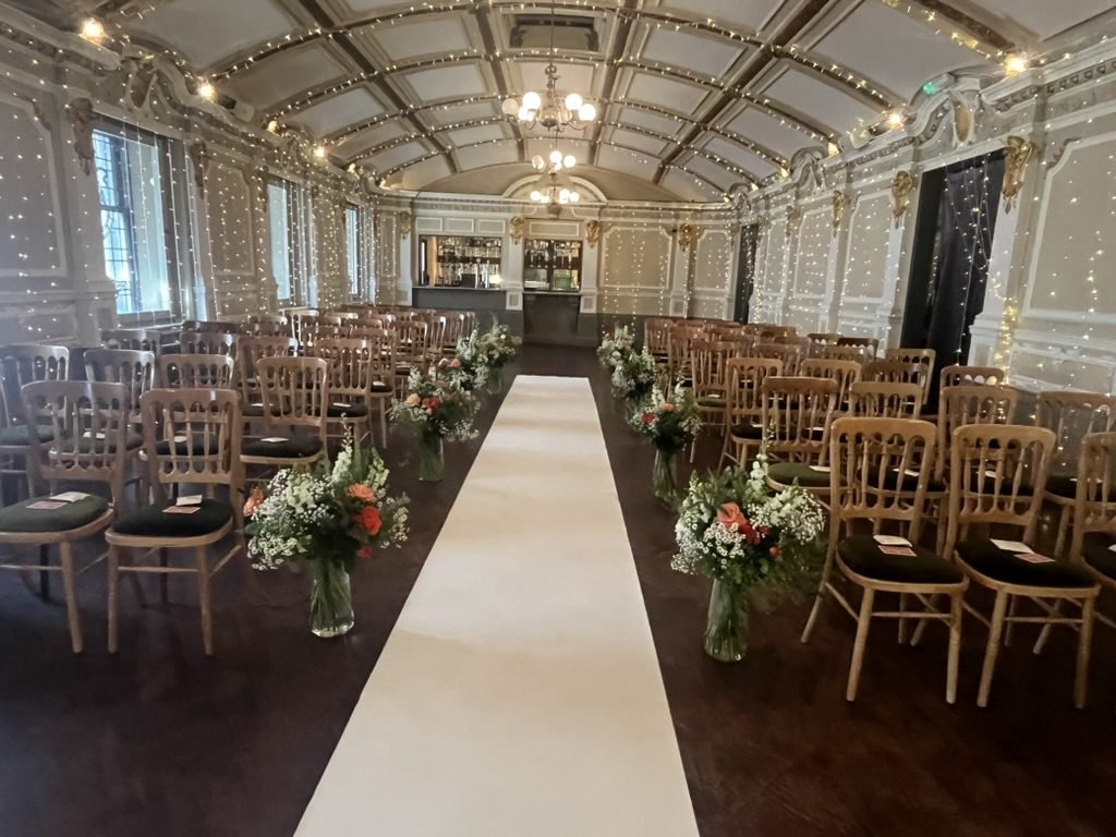 The Grand Ballroom at Sloans set for a humanist wedding, rows of wooden chairs either side of a long white aisle lined with vases of flowers, fairy lights down the walls and chandeliers hanging from the vaulted, coffered ceiling