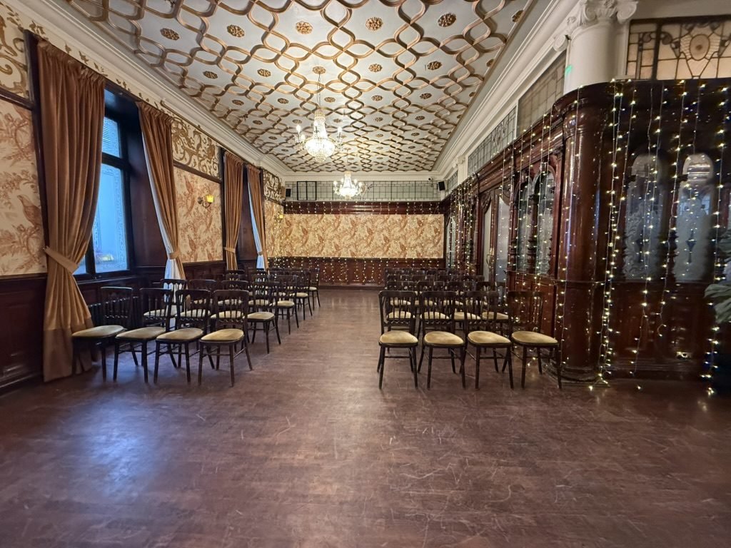 A wood-panelled first-floor dining room at Sloans set with rows of chairs for a winter ceremony: gold patterned walls, a strapwork plaster ceiling, chandeliers, and strings of fairy lights hanging over a carved timber screen with etched glass