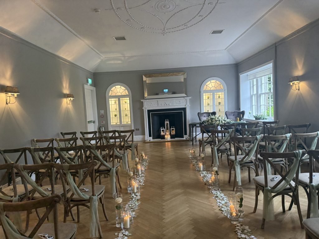 A ceremony room at the Strathaven Hotel set for a humanist wedding in May 2026: wooden cross-back chairs tied with sage ribbon, a herringbone floor, candles and petals lining the aisle, and a white fireplace between arched, glass-fronted alcoves under a decorative plaster ceiling