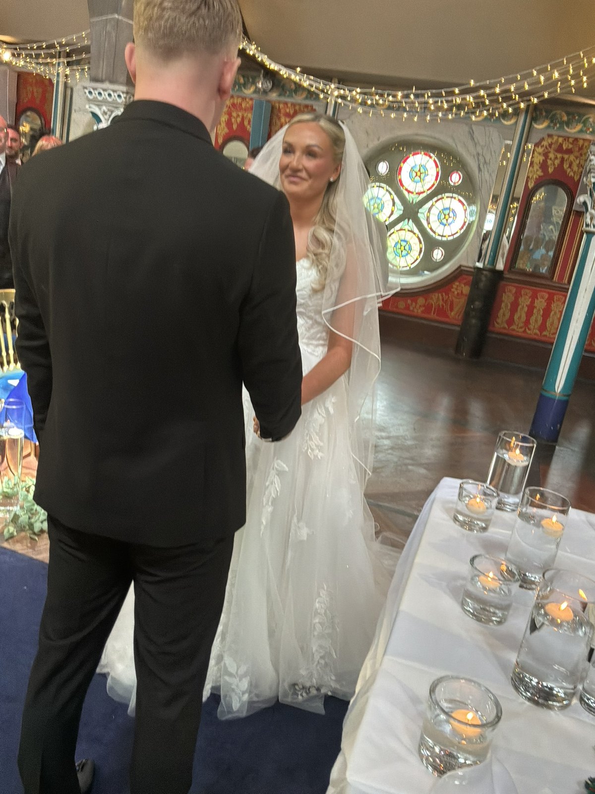 Rebecca smiling at Stuart during their vows at Òran Mór, stained glass and candlelight behind