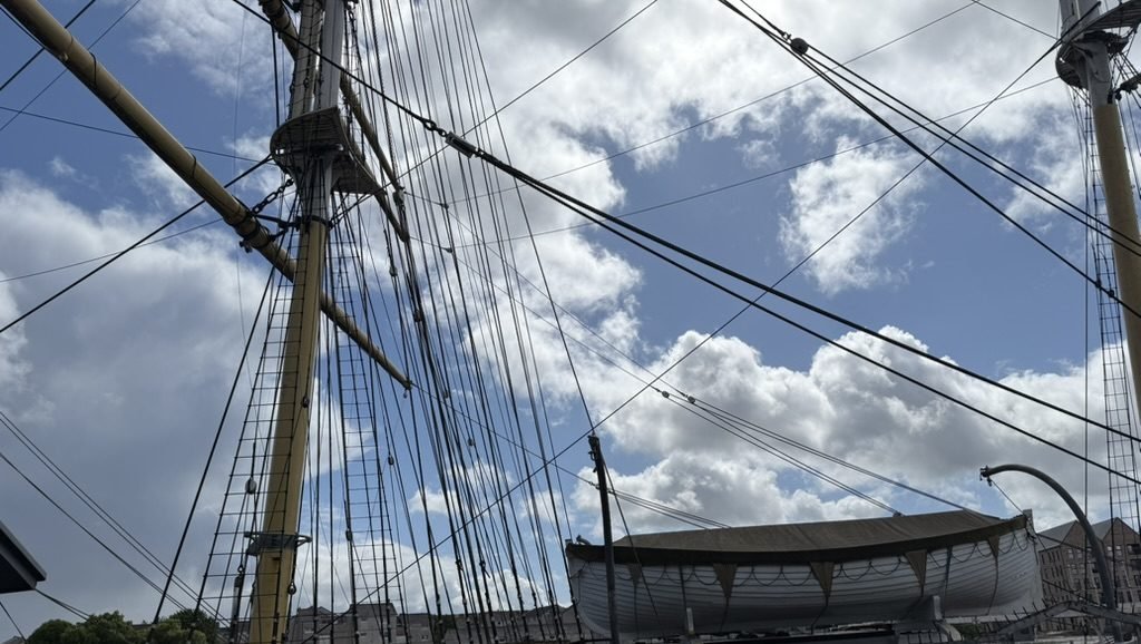 Looking up from the deck of the Tall Ship Glenlee at a yellow mast, its shrouds and rigging, and a white lifeboat against a blue sky full of cloud