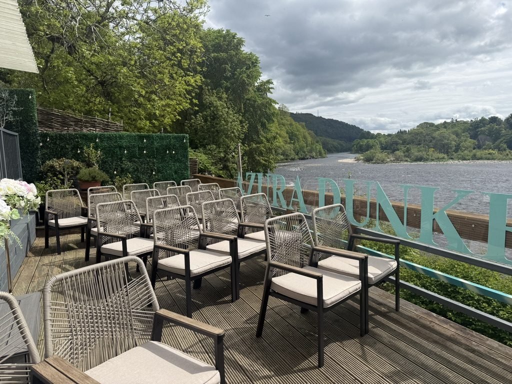 Rows of woven chairs with cream cushions set out on the riverside deck at Dunkeld for a humanist wedding, the wide River Tay and wooded hills beyond, and teal letters along the rail spelling Azura Dunkeld