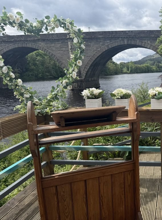 A wooden lectern on the riverside deck in Dunkeld, framed by an arch of white roses, with boxes of white flowers along the rail and the stone arches of Dunkeld Bridge across the River Tay behind