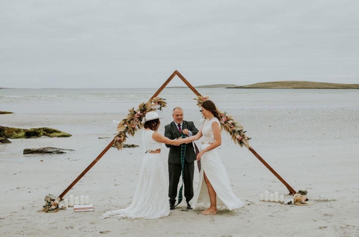 A handfasting during a beach wedding on South Uist — the couple's hands bound in tartan under a flower-dressed arch