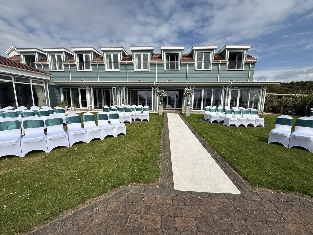 The seaward lawn of the Waterside Hotel set for an outdoor ceremony: rows of white chairs with emerald-green sashes either side of a white runner, leading back to the blue-painted hotel and its glass doors under a summer sky